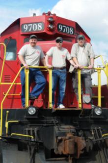 Three men standing on a train car rail.